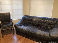 View of the black leather sofa couch alongside the antique leather chair in a room with wooden floor and window blinds.
