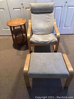 IKEA beige fabric Poang chair with matching ottoman and pine wood three-tiered round side table.