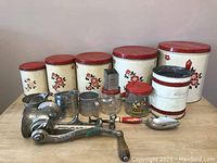Front view of full set showing five cream colored metal baking canisters with red lids and floral designs, plus meat grinder, nutmeg grinder, scoop, sifters, measuring cups, and glass container