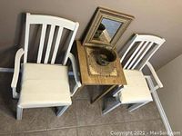 Photo with side table between two white wooden chairs with white cushions. Items placed on table include brass vase, woven placemat, and the framed mirror resting behind.