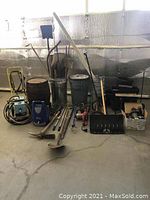 Wide view of assorted garden supplies and tools including tiller, garbage cans, pump, shovels, chainsaw, and tables