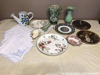Array of vases, plates, napkins, and candy dishes including green vase, white ceramic watering jar, and patterned china plates laid out on beige tablecloth.