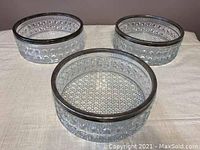 Three large clear crystal bowls with geometric patterned sides and metal silver rims, arranged on a white cloth.