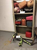 Shelf with several fuel containers, some red metal, some plastic, one blue metal box, and a red metal gas can on floor with metal bike rack.
