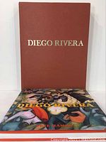Two books displayed, one red hardcover with gold text, the other a colorful dust jacket showing a mural scene by Diego Rivera