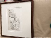 Framed and matted print with visible intricate line drawing of person carrying a child, viewed at an angle on a beige surface.