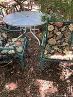 Photo of round glass top patio table and four floral cushioned metal chairs outdoors on wood chips.