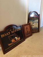 Overall view of three wooden nautical wall hangings arranged against wall: wooden sailboat, Ships Tavern sign, and Boat Repair sign.