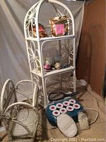 Photo of white wicker floor shelf with three shelves holding bath salts, bath bombs, electric rollers, and ceramic soap pump.