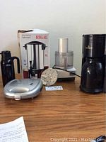 Photo of Cuisinart food processor, Regal coffee maker in box, Gevalia coffee maker, Nissan insulated carafe, and electric sandwich maker on table.