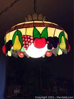 Front view of the Tiffany style leaded stained glass hanging lamp illuminated, showing the fruit pattern of pears, apples, grapes and cherries in multicolor glass.