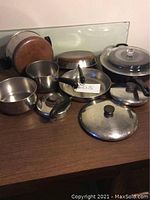 Overview of multiple stainless steel pots and pans with copper bottoms, plastic handles, and various lids on a wooden surface.