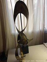 Side and front views of two brass sculptures: sailboat on marble base and abstract brass figure on black pedestal.