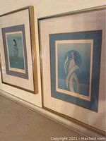 Two gold framed and matted prints leaning against a wall, showing the blue toned stylized figures and double mats.