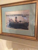 Framed picture showing old man fishing in a boat on water with tables/matters surrounding the image and a gold frame