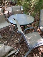 Photo showing round glass top outdoor table and four outdoor chairs around it, two in dark grey and two in light grey, on ground with leaves and shaded by trees.