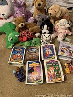 Photo showing the collection of plush toys, Disney VHS tapes, and McDonald's figures spread on a carpet. Plush animals include bears, dogs, a koala, frog, ladybug, turtle, and lamb. VHS tapes are arranged in front.