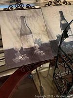 Two decorative pictures with wine bottle and glass artwork framed with black metal scrollwork on top and bottom, visible with parts of black metal wine rack