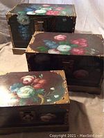 Front view of three wooden storage boxes of varying sizes with painted floral designs and metal corner accents, stacked side by side.
