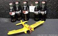 Photo showing five Coca-Cola Classic Santa Claus 1994 commemorative glass bottles in a six-pack holder with a yellow Northrop Gamma die-cast metal bank airplane in front.