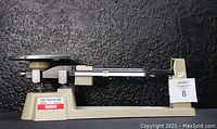 Side view of the beige OHAUS triple beam balance scale with round metal pan and sliding weights visible.