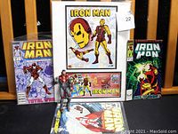 Full view of the lot showing the matted art in a black frame, comic books, and the Iron Man action figure posed on the chair.