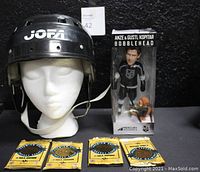 Jofa black LA Kings hockey helmet displayed on styrofoam head, sealed Leaf NHL card packs arranged in a row in front, Anze & Gustl Kopitar bobblehead boxed behind them.