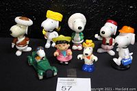 Seven Peanuts figures assorted with a green cartoon car and Snoopy driver; mainly Snoopy and one Lucy figure in varied costumes