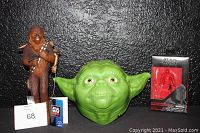 Three Star Wars collectibles shown against black background: Chewbacca figure standing with rifle, green 3D light-up Yoda face mask wall mount, and boxed Elite Praetorian Guard figure.