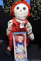 Large vintage Raggedy Ann plush doll with red yarn hair wearing red dress and floral blouse, standing behind boxed new old stock smaller Raggedy Ann doll.