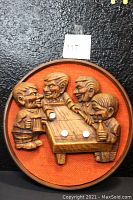 Front view of the circular mid-century modern wall mounted 3D art showing four men playing a skeet board game around a table with drinks in hand and game pieces on the table, set against an orange fabric background.