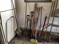 Wide shot showing multiple garden and construction tools including rakes, broom, spades, crowbar, axe, and handles against a wall.
