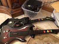 Two black Rock Band guitar controllers with colored fret buttons and attached straps, laid on carpet next to plastic bin full of cables and accessories.