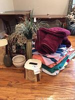 Photo showing stack of assorted colorful towels and linens with artificial floral arrangement in tall ceramic vase and other decorative items on hardwood floor