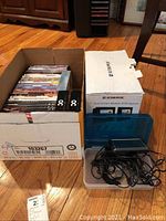 Box containing DVDs and boxed Audiovox dual screen mobile DVD system on wooden floor near wooden furniture.