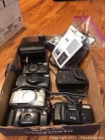 Box containing various vintage cameras, camera cases, instruction manuals, and cables.