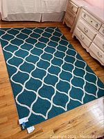 Top view of teal area rug with sand lattice pattern on wood floor, beside white bedroom furniture.