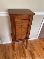 Front view of medium-tone wooden jewelry cabinet with five drawers and side doors, standing on four legs.