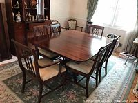 Wide shot of dining table set with six chairs on floral rug in a room with wooden furniture and patterned curtains.