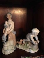 Two porcelain figurines on dark wood shelf, girl holding dog and boy with dog.