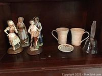 Full view of 4 figurines, 2 Belleek cups, decorative plate, and glass bell arranged on wooden shelving.