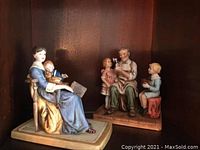 Photo of two Norman Rockwell inspired figurines placed in a wooden shelf, showing a woman in a blue dress holding a child and a book, and an elderly man with two children seated nearby.