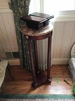 Full view of marble top display stand with dark wood legs and bottom base, standing near a window and curtain.