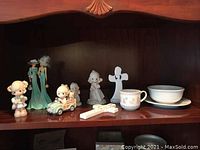 Shelf display of assorted Precious Moments figurines, baby cup, bowl, and plate with pastel floral designs, arranged together indicating the full lot contents