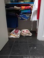 Folded assorted tablecloths on shelf with stacks of placemats and dishtowels on floor