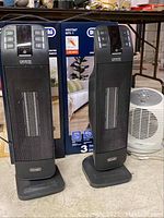 Two DeLonghi black tower heaters and smaller white Patton oscillating heater all placed on floor with original boxes in background.