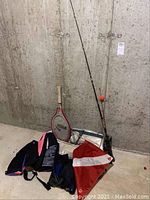 Fishing rods, tennis racket, scuba flag, tennis bag on concrete floor against wall