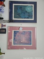 Two framed wall art prints hung on a white wall, one with a dark blue mat and the other with a pink mat.