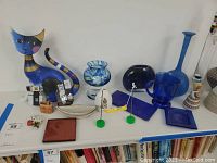 Photo of a white table with a large artistic cat figurine, blue glass vases, small figurines, trays, and a Galileo thermometer.