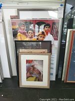 Five framed wall art pieces stacked against a wall showing various subjects including group portraits and elderly man with turban